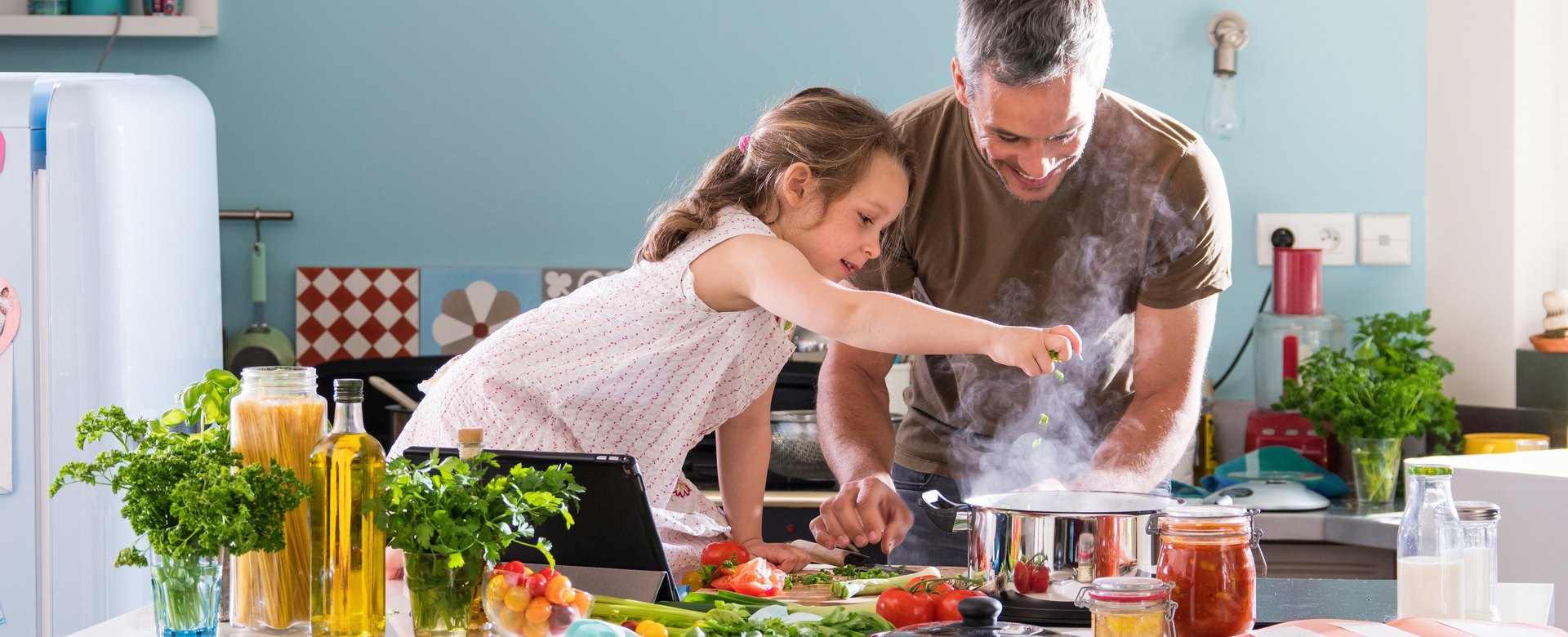 Vater und Tochter kochen gemeinsam ein Gericht mit Nudeln, Olivenöl und Tomatensugo.
