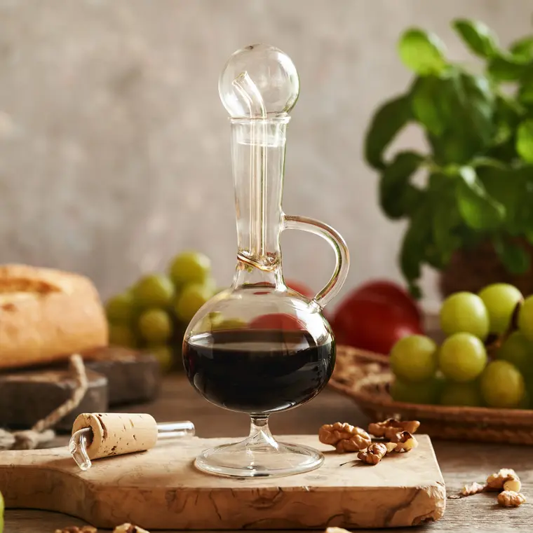 Eine Glaskaraffe gefüllt mit Balsamico-Essig auf einem Holzbrett, umgeben von grünen Trauben, Tomaten, Basilikum, Nüssen und Ciabatta