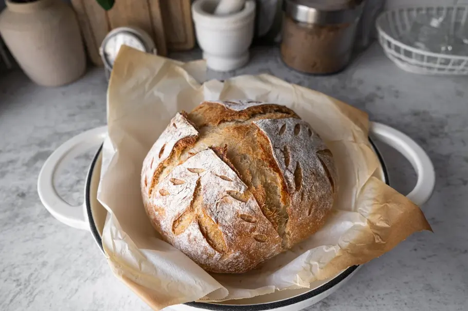 Ein Brotlaib aus Sauerteig auf Backpapier in einer weißen Porzellanschüssel, im Hintergrund Gewürze