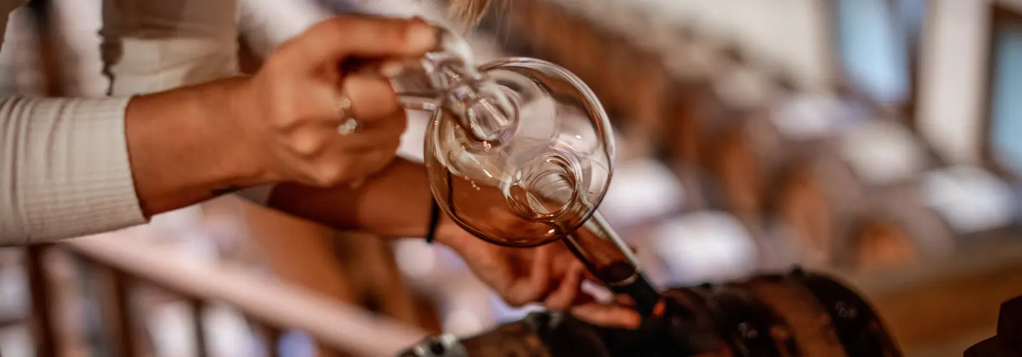 Nahaufnahme einer Frau, die Balsamico-Essig mit einem Glasinstrument aus einem Holzfass entnimmt. 