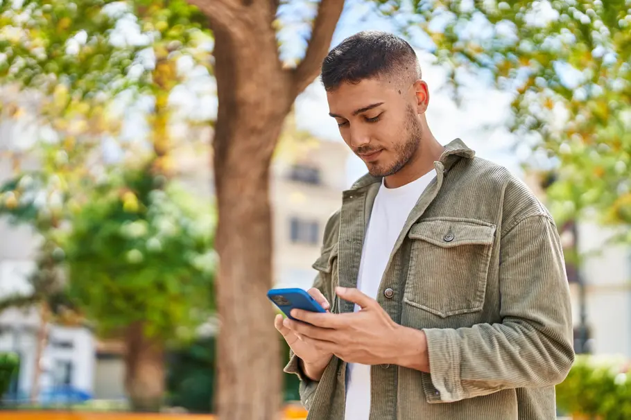 Ein junger Mann in weißem T-Shirt und grünem Hemd blickt auf sein Smartphone in einem Park.