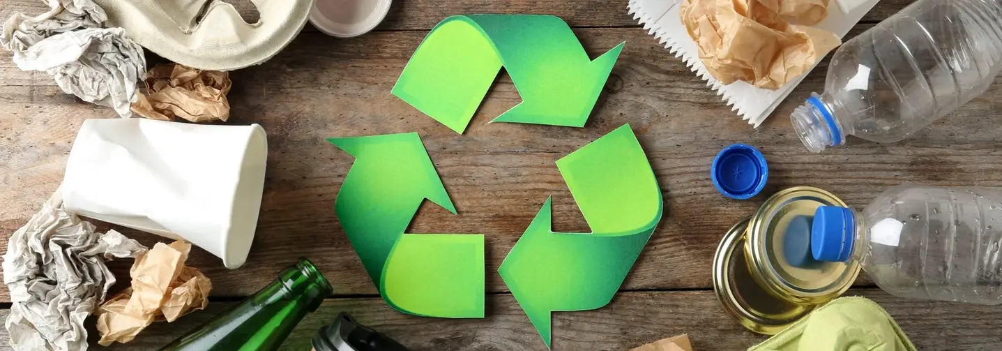 Grünes Recycling-Symbol auf Holzuntergrund, umgeben von verschiedenen Verpackungen, etwa Glas- und PET-Flaschen, Kunststoffbecher, Coffee to go Becher, Eierkartons.
