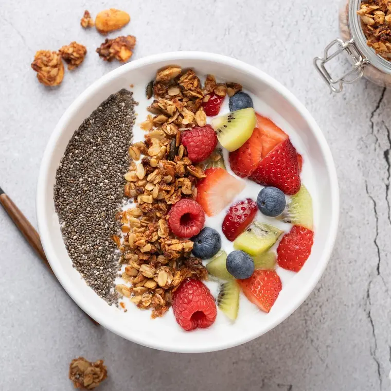 Draufsicht auf eine weiße Schale mit Kefir, Müsli, Chiasamen und frischen Früchten. Links ein Metalllöffel; rechts ein offenes Müsliglas und eine Schale mit Obst.
