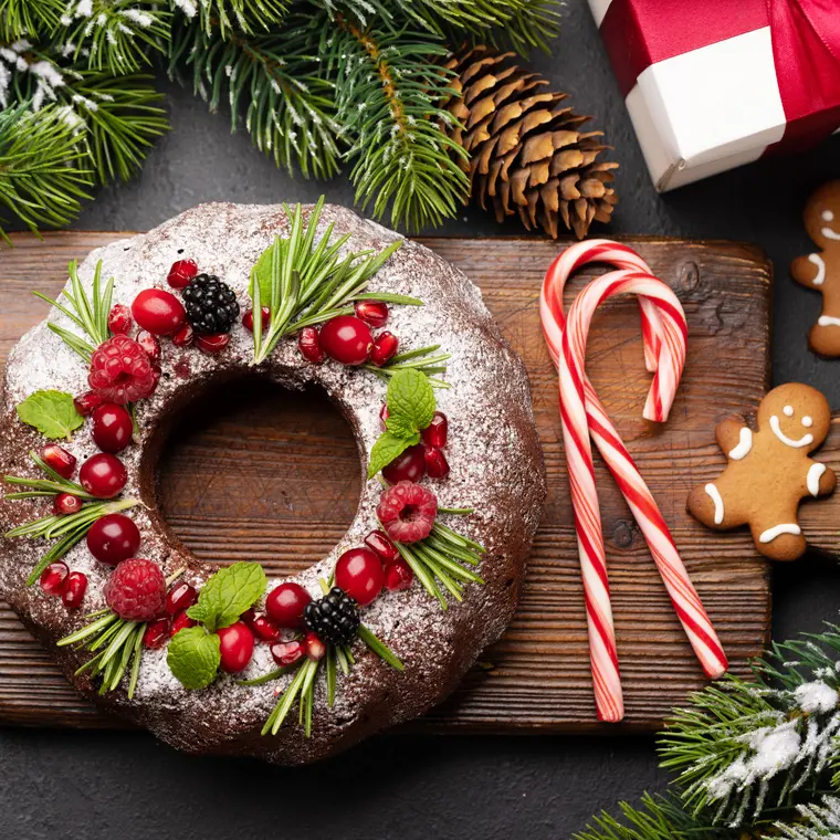 Weihnachtlich dekorierter Gugelhupf auf einem Holzbrett, umgeben von Zuckerstangen, Lebkuchen und Tannenzweigen.