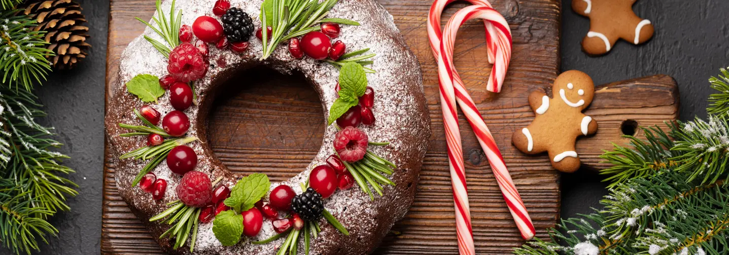 Weihnachtlich dekorierter Gugelhupf auf einem Holzbrett, umgeben von Zuckerstangen, Lebkuchen und Tannenzweigen.