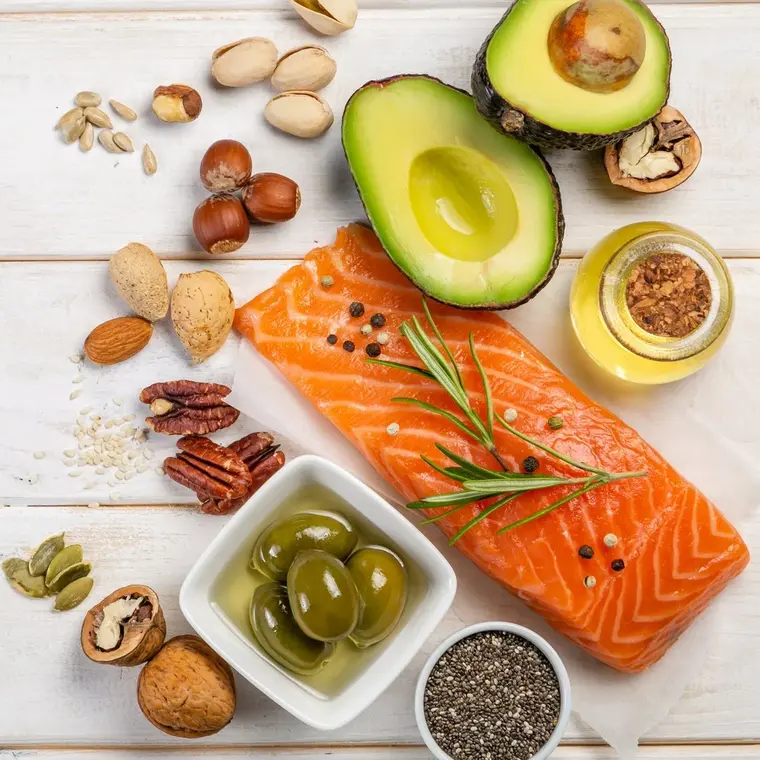 Verschiedene Omega-3 haltige Lebensmittel in Draufsicht auf einem weißen Holztisch, unter anderem Lachs, Olivenöl, Oliven, verschiedene Nüsse und Samen sowie eine halbe Avocado.