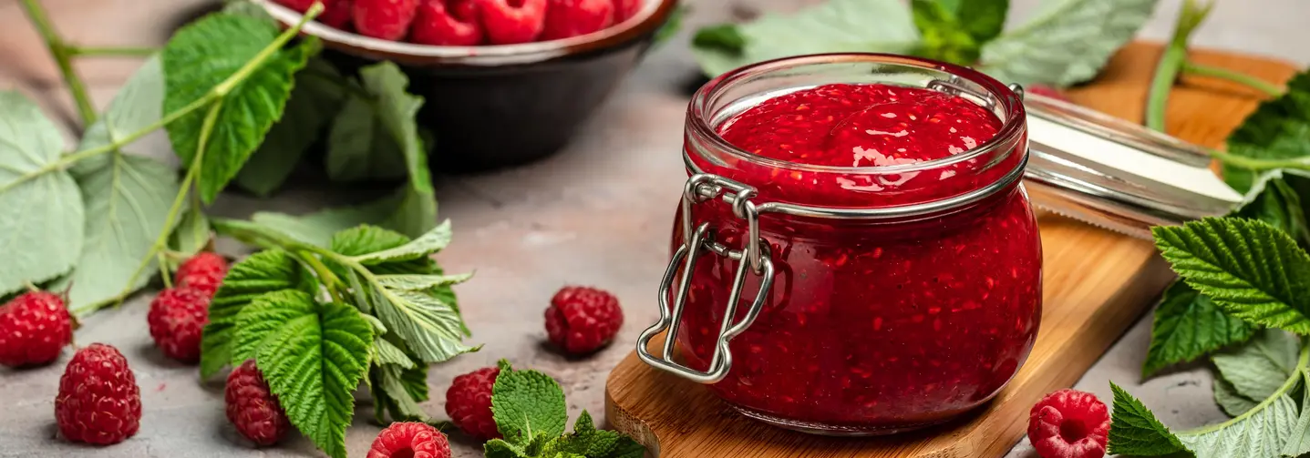 Einmachglas mit Himbeermarmelade auf einem Holzbrett, umgeben von frischen Himbeeren und grünen Blättern.