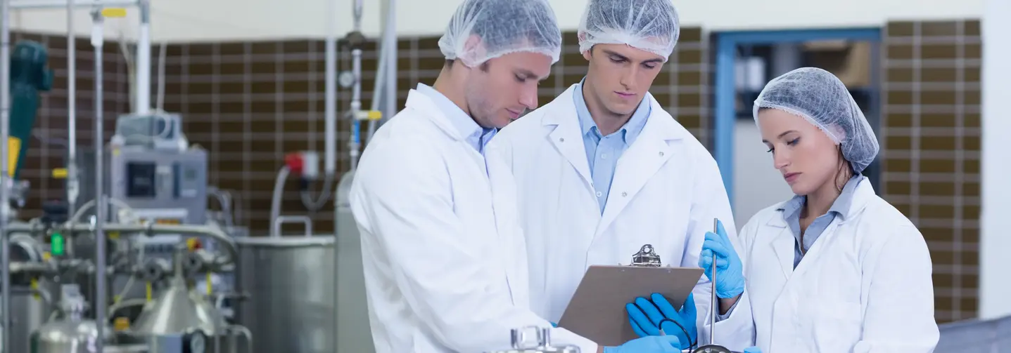 Zwei Männer und eine Frau in weißen Arbeitsmänteln, mit transparentem Kopfschutz und blauen Handschuhen vor einer Produktionsanlage.