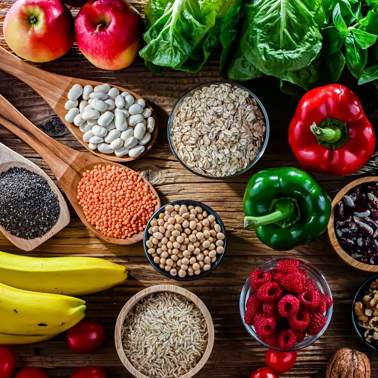 Ballaststoffreiche Lebensmittel von oben auf einem Holztisch – unter anderem Bananen, Beeren und Äpfel, Schälchen mit verschiedenen Samen, Bohnen und Nüsse.