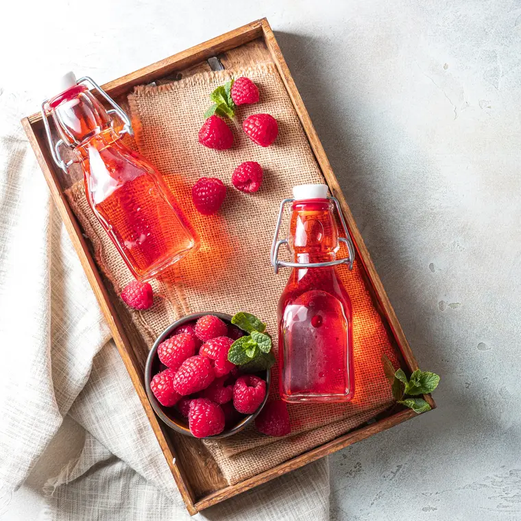 Ein Holztablett auf Marmoruntergrund. Darauf zwei Glasflaschen gefüllt mit Himbeeressig, umgeben von frischen Himbeeren. 