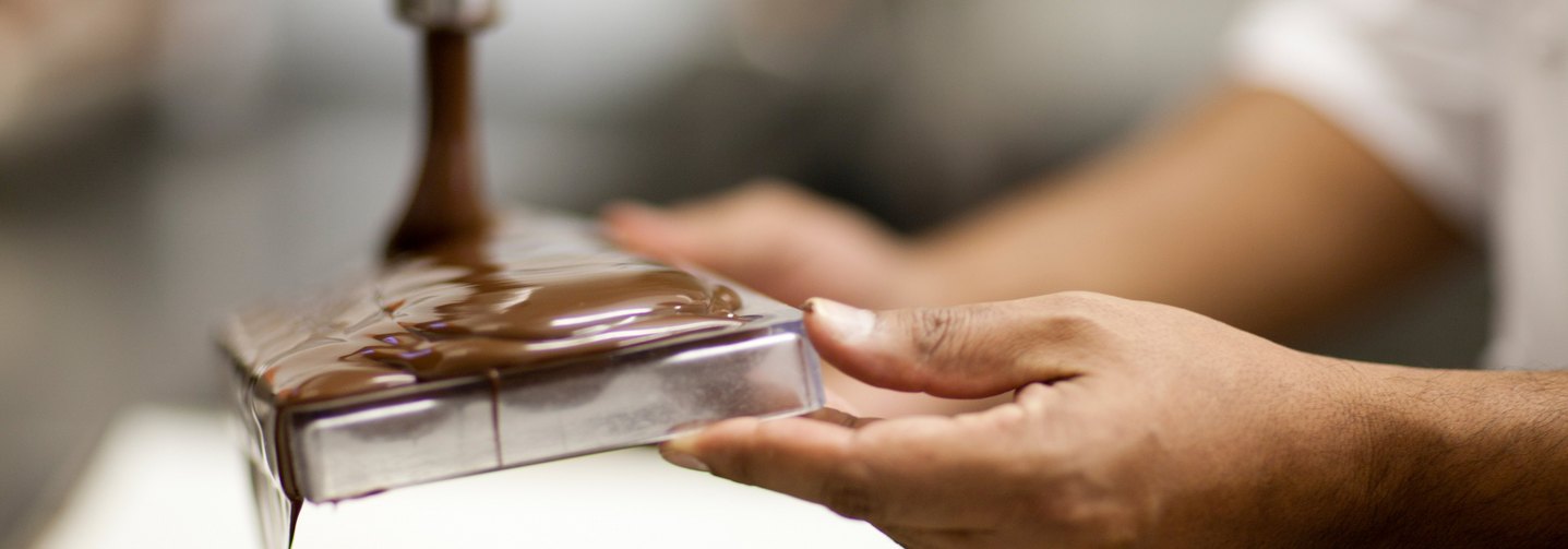 Schokoladen und Schokoladenerzeugnisse - hier flüssige Schokolade, die aus einem Rohr kommt - werden je nach Rezeptur aus Kakaobutter, Kakaomasse, Kakaopulver und Zucker hergestellt.