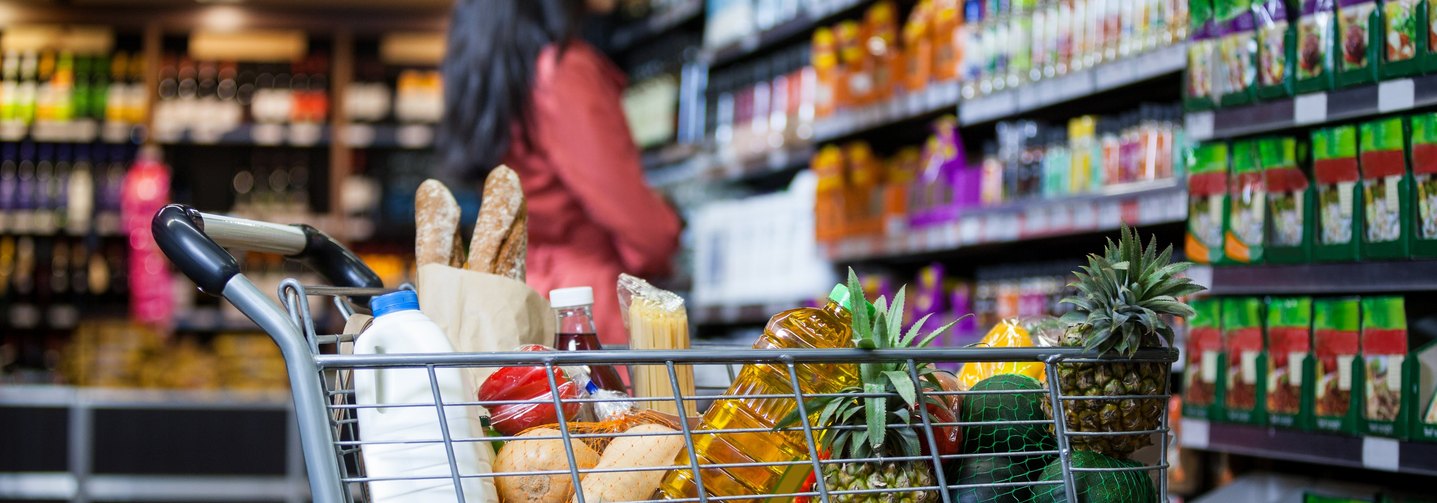 Eine Frau mit vollem Einkaufswagen vor einem gut gefüllten Supermarkt-Regal.