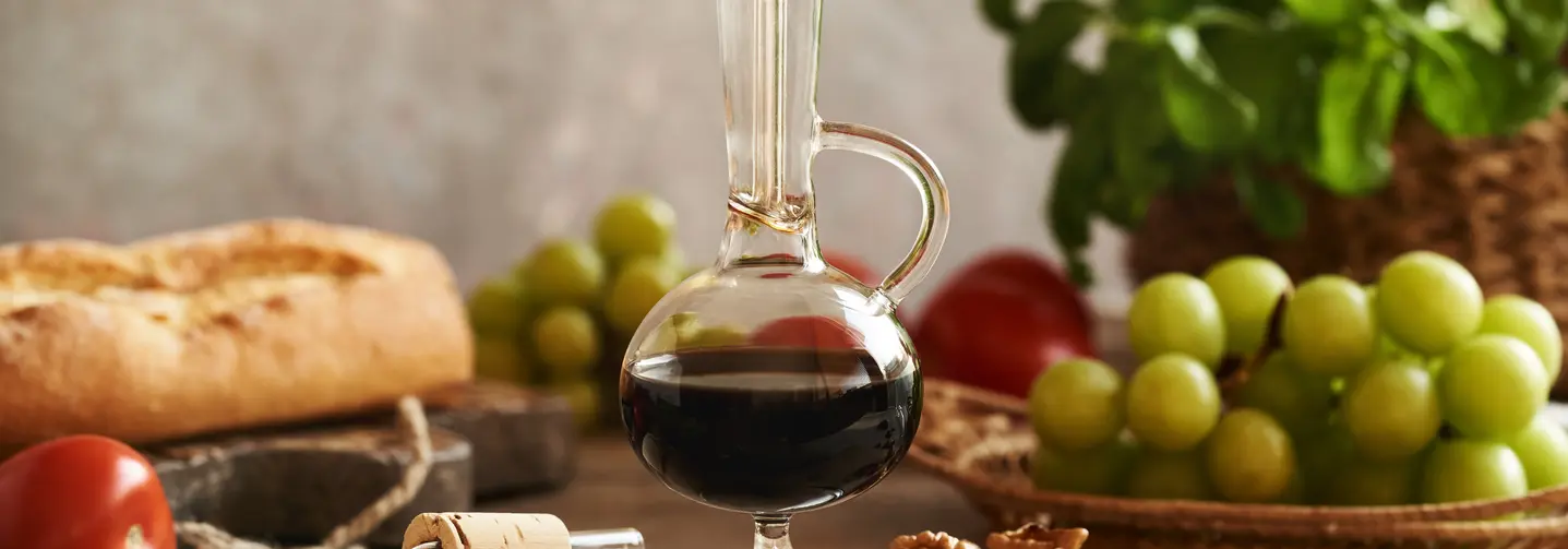 Eine Glaskaraffe gefüllt mit Balsamico-Essig auf einem Holzbrett, umgeben von grünen Trauben, Tomaten, Basilikum, Nüssen und Ciabatta. 