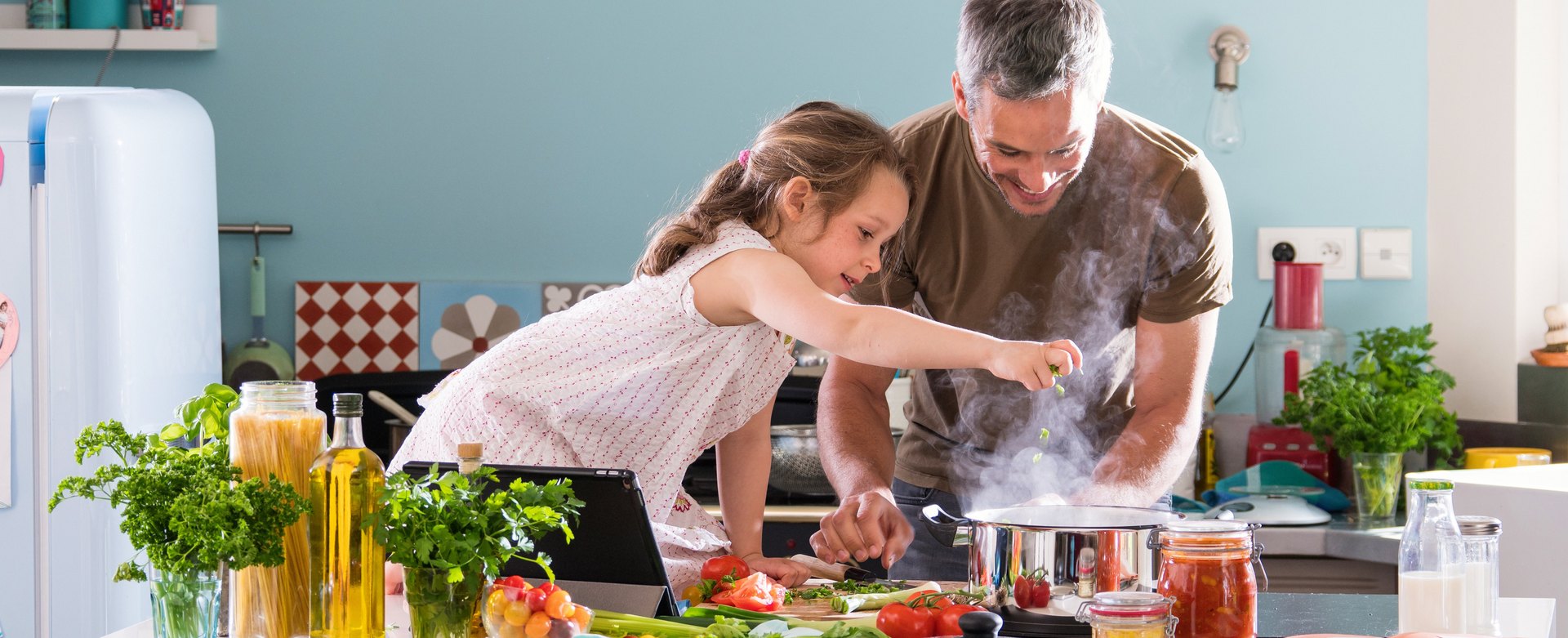 Vater und Tochter kochen gemeinsam ein Gericht mit Nudeln, Olivenöl und Tomatensugo.
