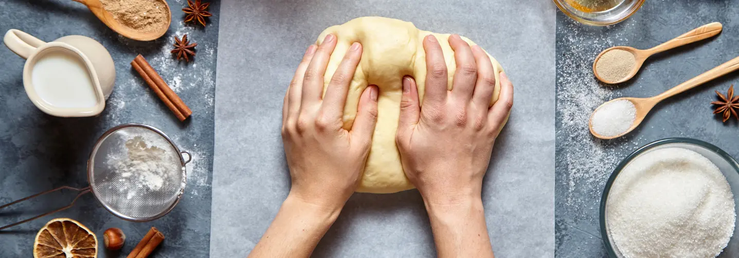 Zwei Hände, ein Kuchenteig und verschiedene Zutaten wie Zucker, Eier und Mehl von oben.