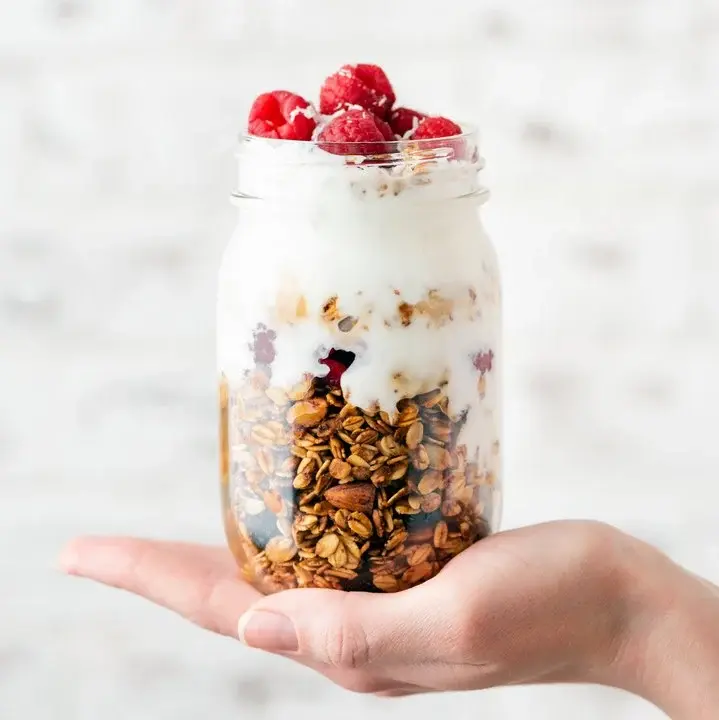 Himbeer-Frucht-Dessert mit Skyr und Granola in ein Weckglas geschichtet, das von einer Hand gehalten wird.