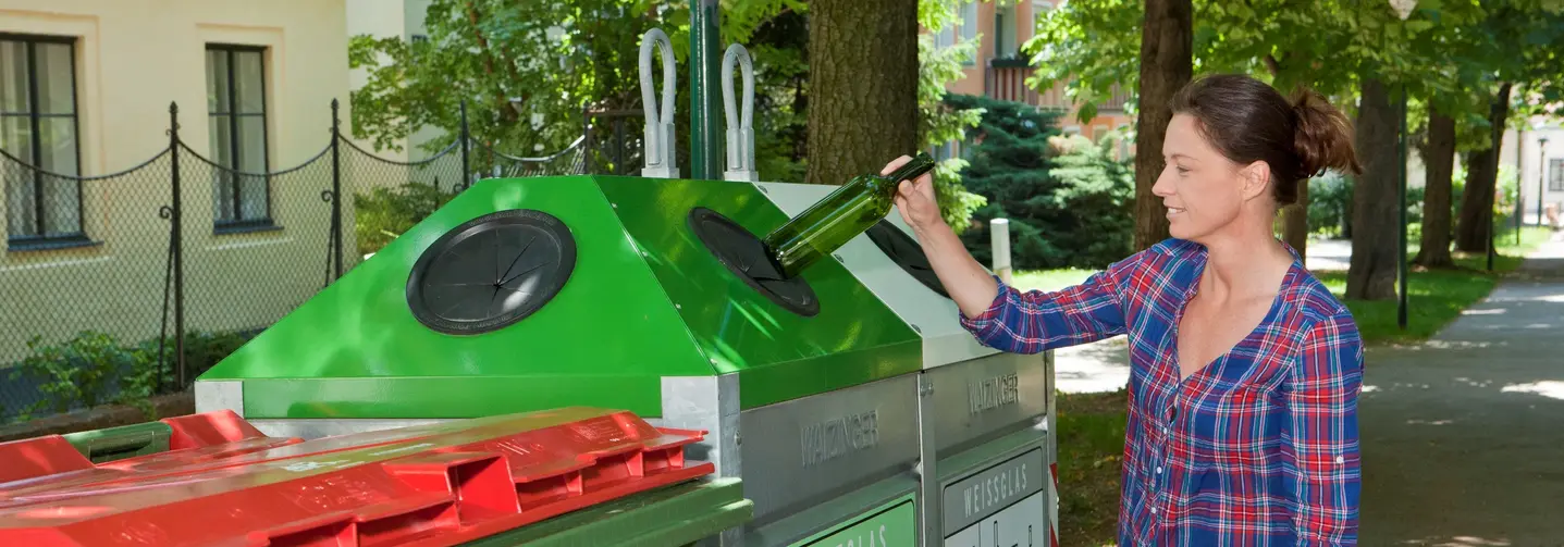 Eine Frau in einer gemusterten Bluse wirft eine Buntglasflasche in den Altglascontainer bei einer Sammelinsel. 