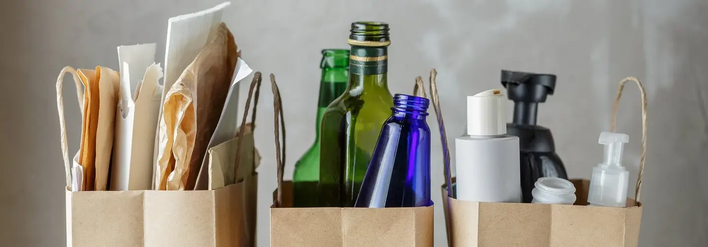 Drei Papiertüten befüllt mit verschiedenen Verpackungsmaterialien: Papier, Glas und Kunststoff.