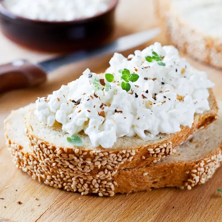 Zwei Brotscheiben auf einem Holzbrett, mit Cottage Cheese (Hüttenkäse) bestrichen.