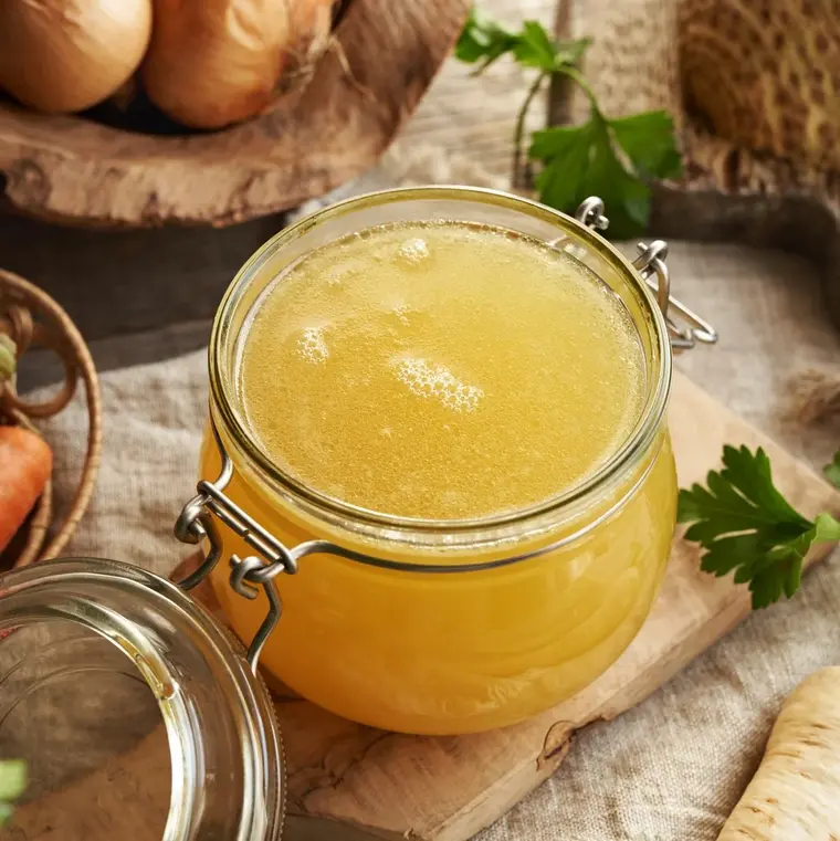 Einmachglas mit Knochenbrühe (Bone Broth) auf einem Tisch, daneben Wurzelgemüse wie Karotten und Sellerie.