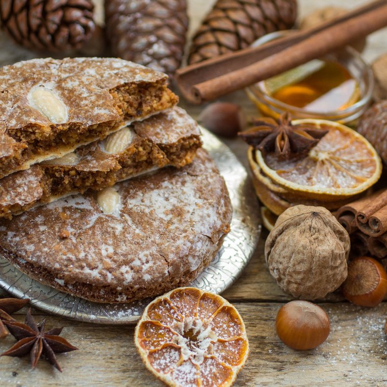 Oblatenlebkuchen mit Zuckerguss umgeben von Zimtstangen, Gewürznelken und Nüssen auf einer Holzfläche.