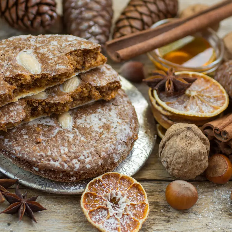 Oblatenlebkuchen mit Zuckerguss umgeben von Zimtstangen, Gewürznelken und Nüssen auf einer Holzfläche.