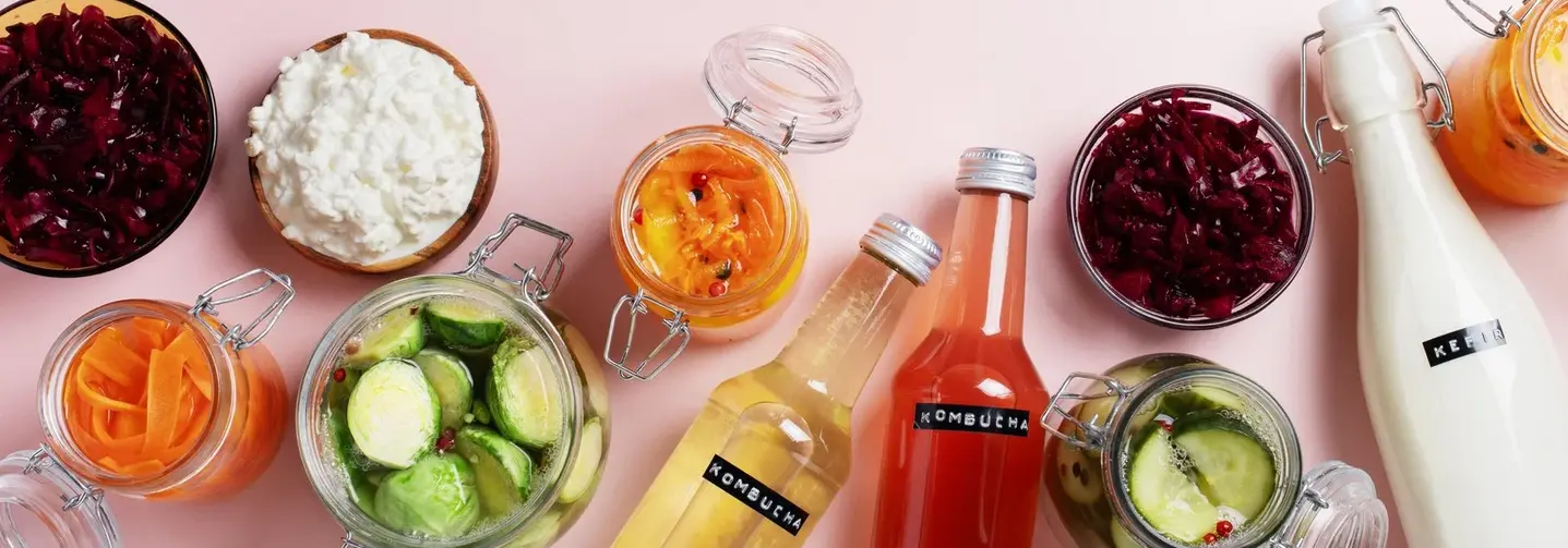 Verschiedene fermentierte Lebensmittel in Gläsern, Flaschen und Schälchen auf rosa Untergrund – unter anderem Sauerkraut, Rotkraut, Salzgurken, Hüttenkäse, Kefir, Kombucha und eingelegtes Gemüse