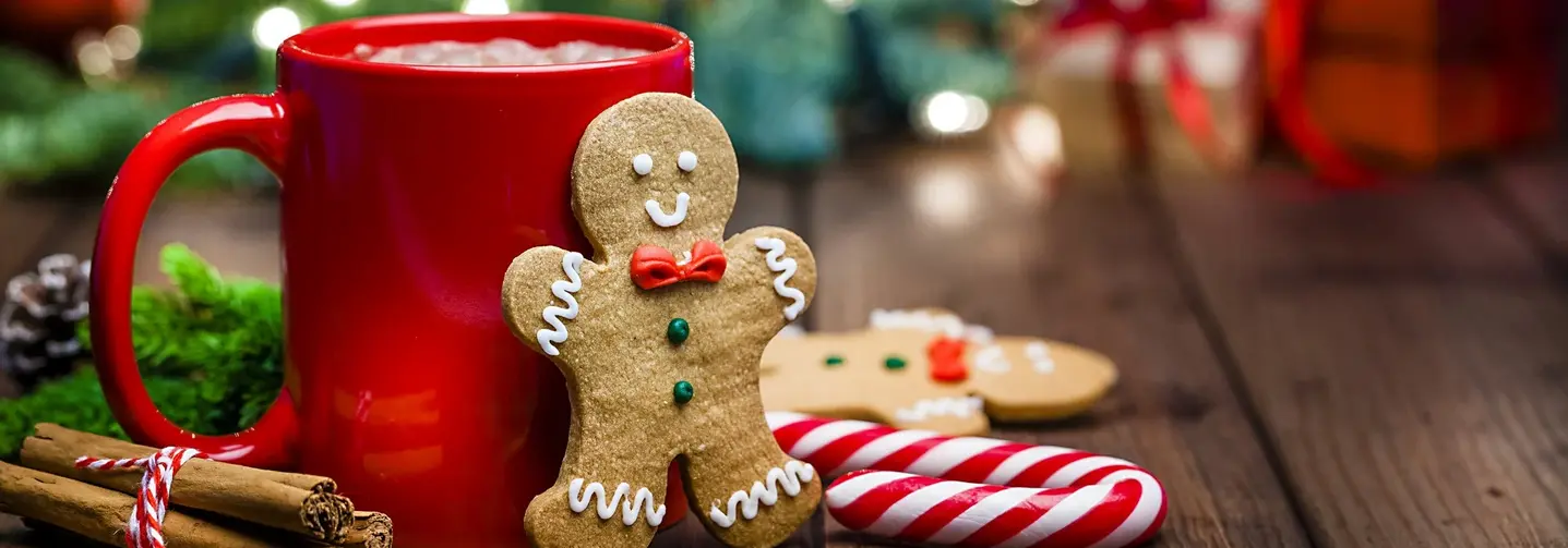 Lebkuchenmännchen, eine rote Tasse mit heißer Schokolade, eine Zuckerstange und Zimtstangen vor weihnachtlichem Hintergrund